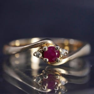May include: A gold ring featuring a central round-cut deep red ruby held in a four-prong setting, flanked by two small clear accent stones on either side. The band has a smooth, polished finish and a bypass design that curves around the center stones.