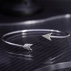 May include: A silver-coloured arrow bracelet. The arrowheads are at each end of the open cuff. One arrowhead is detailed with small, sparkling stones. The bracelet is displayed on a reflective surface.