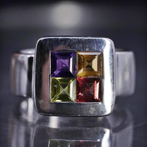 May include: A silver ring with a square setting, showcasing four square gemstones in purple, yellow, green, and red. The ring has a chunky band and a reflective surface.