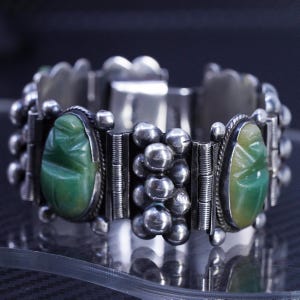 Peut inclure: Un bracelet à maillons en argent orné de deux pierres ovales vertes sculptées en forme de scarabée. Le bracelet est composé de sections alternées de sphères en argent poli et de barres verticales, reliées par des charnières.