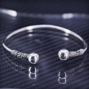 May include: A silver bangle bracelet with two round balls on the ends. The bracelet has a textured design.