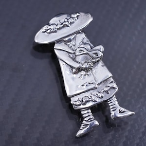 Puede incluir: Un broche de plata que representa a una mujer con un sombrero de ala ancha y sosteniendo un animal pequeño en sus brazos.