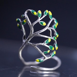 May include: A silver-toned open ring featuring a branching tree design. The branches are adorned with small, oval-shaped leaves finished in vibrant green enamel with bright yellow centers, creating a detailed botanical motif on the metal band.