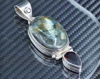 Puede incluir: Un colgante de plata con una gran piedra de labradorita de forma ovalada, con un brillo iridiscente verde oscuro y azul. El colgante está adornado con dos pequeñas piedras de granate en forma de lágrima.
