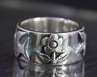 Puede incluir: Anillo plateado con diseño floral. El anillo presenta flores y hojas en relieve alrededor de la banda, con un color de acento azul claro. El anillo es reflectante, con un fondo oscuro.