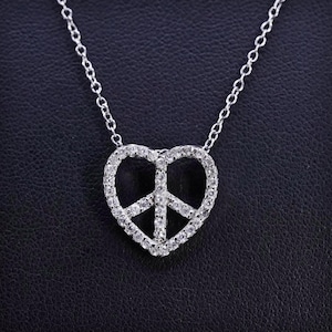 May include: A silver necklace featuring a heart-shaped pendant with a peace sign design. The pendant is encrusted with small, clear gemstones, and the chain is delicate. The necklace is displayed against a dark background.