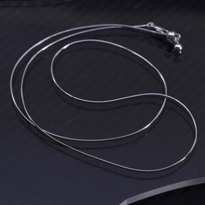 May include: A delicate silver necklace with a thin, flat chain. The necklace is displayed on a dark surface, showcasing its simple elegance. The clasp and end details are visible, highlighting the craftsmanship of the jewelry.