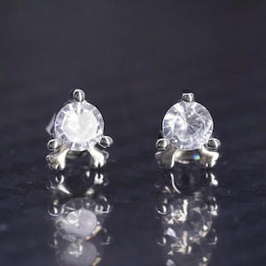 May include: A pair of silver stud earrings with clear round cubic zirconia stones. The stones are set in a four-prong setting and the earrings have a small, delicate design.