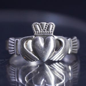 May include: A silver Claddagh ring featuring a central heart held by two hands, topped with a crown. The metal has a polished finish with visible detailing on the cuffs and the crown. The design is symmetrical, showcasing the traditional motif.