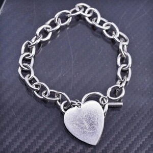 May include: Silver-toned bracelet featuring a heart-shaped charm. The charm is connected to a chain-link bracelet with a toggle clasp. The heart charm has a slightly textured surface.