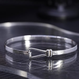 May include: A silver-colored bangle bracelet with a hook and eye closure. The bracelet is round and has a simple, elegant design. The hook closure is a rectangular shape. The bracelet is displayed on a reflective surface.