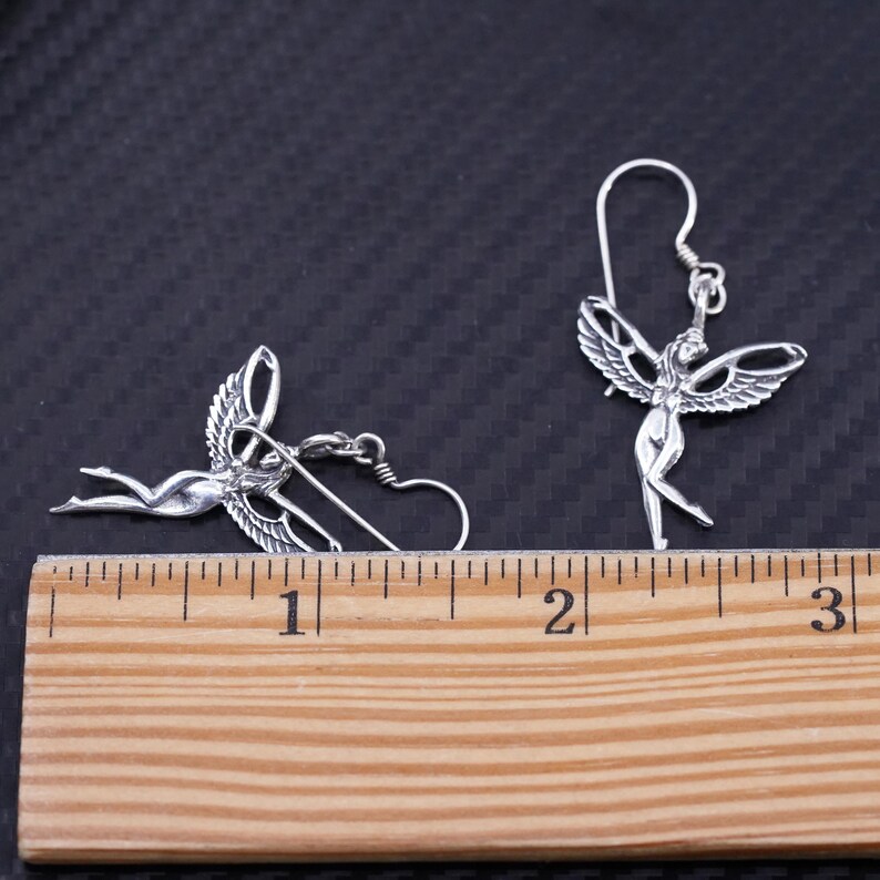 K&ouml;nnte beinhalten: Ein Paar silberne Ohrringe mit einem gefl&uuml;gelten Figuren-Design. Die Ohrringe sind neben einem Lineal abgebildet, wobei jeder Ohrring etwa 2,5 cm lang ist.