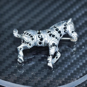 Puede incluir: Broche de cebra plateada con detalles de piedras negras. El broche presenta un diseño detallado de un cebra con piedras negras incrustadas en sus rayas. El broche tiene un alfiler en la parte posterior.