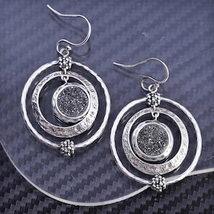 May include: A pair of silver-toned dangle earrings. Each earring features three concentric circles, with a textured outer ring and a central circle filled with dark, sparkling material. The earrings have a hook closure.