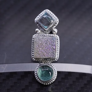 May include: A silver pendant with three gemstone settings. The top setting is a square-cut, blue-green stone. The middle setting is a square, iridescent stone. The bottom setting is a round, green stone.