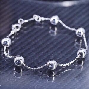 May include: A silver chain bracelet with round silver beads. The bracelet is on a black textured surface.