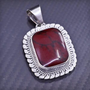 May include: A silver pendant with a red and black stone set in a silver frame. The frame has a decorative border.