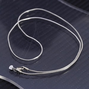 Könnte beinhalten: Eine silberfarbene Schlangenkette mit einem herzförmigen Anhänger in der Nähe des Verschlusses. Die Halskette besteht aus einer feinen, flexiblen Kette und wird auf einer dunklen Oberfläche präsentiert. Der Schmuck ist für den täglichen Gebrauch bestimmt.