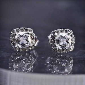 Puede incluir: Un par de pendientes de botón plateados con una piedra central cuadrada y transparente. Cada pendiente está enmarcado por un halo de piedras redondas más pequeñas. Los pendientes se presentan sobre una superficie reflectante.