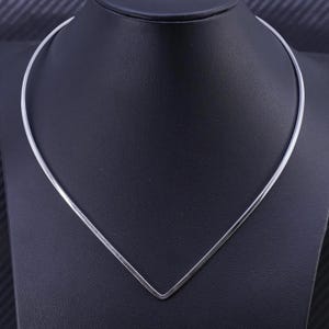 Könnte beinhalten: Eine minimalistische silberfarbene Halskette mit einem V-förmigen Anhänger. Die Halskette wird auf einer schwarzen Oberfläche präsentiert, was ihr schlankes Design hervorhebt. Das Schmuckstück ist ein schlichtes, elegantes Accessoire.