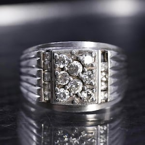 May include: A silver ring with a rectangular face set with multiple round, clear gemstones. The band has a ridged design on the sides. The ring is displayed on a reflective surface.