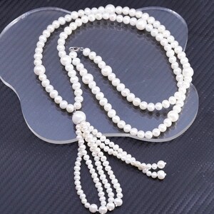 May include: A white pearl necklace with a tassel pendant. The necklace features multiple strands of small, round pearls and a larger pearl at the tassel's base. The necklace is displayed on a clear, wavy surface.