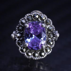 May include: A silver ring featuring a central oval-cut purple gemstone held by four prongs. The stone is surrounded by a scalloped border of small, dark, faceted marcasite stones set in a metallic frame with a textured, darkened finish.