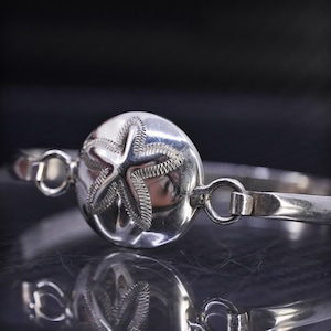 May include: A silver bracelet featuring a large, round centerpiece with a detailed starfish design. The bracelet has a simple, elegant design with a clasp closure. The jewelry is reflective and appears to be made of polished metal.