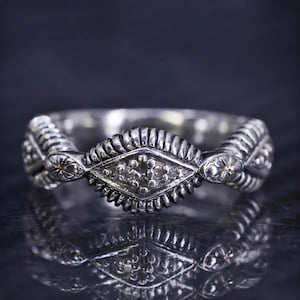 Peut inclure: Une bague en argent avec une pierre centrale en forme de diamant entourée de diamants plus petits. La bague a un design détaillé et orné.