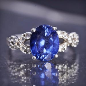 May include: A silver ring featuring a large, oval-cut, deep blue gemstone as the focal point. The band is adorned with smaller, clear gemstones, creating a sparkling contrast. The ring's design is elegant and eye-catching, suitable for special occasions.