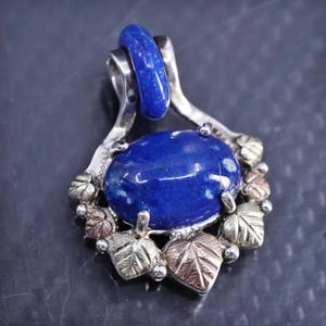 May include: A silver pendant with a large, oval, deep blue stone. The stone is surrounded by a decorative frame with gold and silver leaf accents. A smaller blue stone is at the top of the pendant.