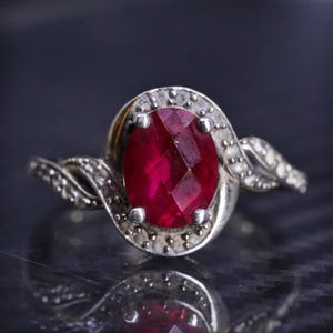 May include: A silver ring with a large, oval-shaped ruby gemstone surrounded by smaller diamonds. The ring has a twisted band.