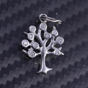 Könnte beinhalten: Ein silberner Baum-Anhänger mit kleinen, runden, klaren Edelsteinen, die die Blätter darstellen. Der Anhänger hat eine Öse zum Anbringen an einer Kette. Das Baumdesign ist detailliert, und die Edelsteine sind in einer Fassung gefasst.