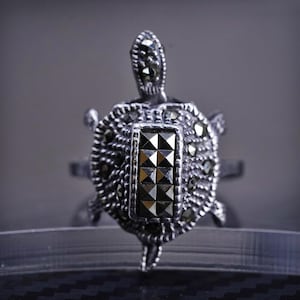 May include: A silver turtle ring with a black marcasite stone in the center. The turtle is detailed with intricate designs and is a statement piece.