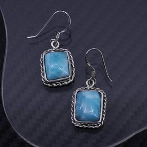 May include: A pair of silver earrings featuring rectangular, light blue gemstones. Each stone is set within a twisted rope-like border and hangs from a simple hook. The earrings have a polished, reflective surface.