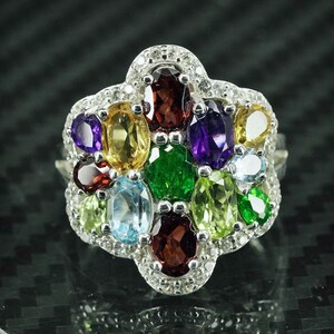 May include: A silver ring with a cluster of colorful gemstones, including green, red, purple, blue, and yellow. The ring is set in a flower-shaped design.