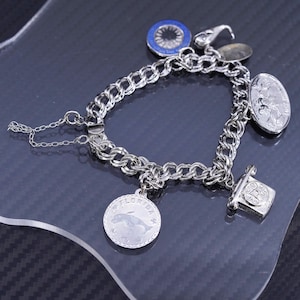 May include: A silver charm bracelet with various charms, including a dolphin, a coin, and a blue and white emblem. The bracelet is made of a thick chain and has a clasp closure. The charms are detailed and add visual interest.