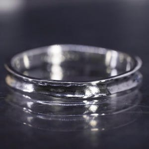 May include: A silver ring with a textured, hammered finish. The ring has a simple, elegant design with a slightly raised outer edge. The ring is reflective, with a dark background.