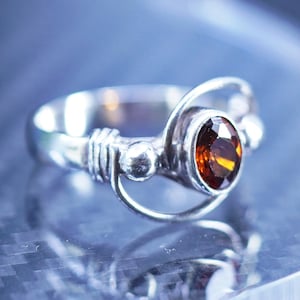 May include: A silver ring with a brown gemstone set in a unique, abstract design. The ring features a twisted band and a small ball accent.