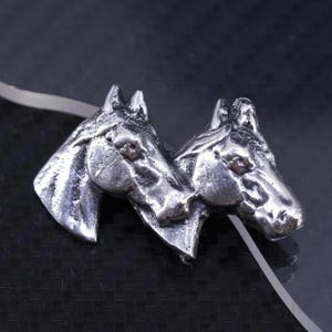 Puede incluir: Una escultura de metal plateado que representa dos cabezas de caballo juntas. Los caballos muestran detalles de crines y rasgos faciales. El conjunto se encuentra sobre un fondo oscuro.