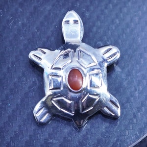 May include: A silver turtle pendant featuring a red gemstone set in the centre. The turtle's shell is decorated with a geometric design.