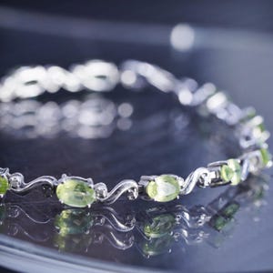May include: A silver-toned metal bracelet featuring a series of oval-cut, light green peridot gemstones. The stones are set in individual prongs and connected by curved, S-shaped silver metal links that create a continuous, flowing design.