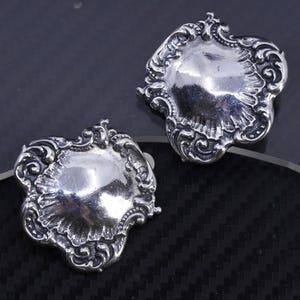 May include: Two ornate, silver-toned metal earrings. Each earring features a central, slightly domed design surrounded by intricate scrollwork and a scalloped edge. The earrings are displayed on a dark, textured surface.