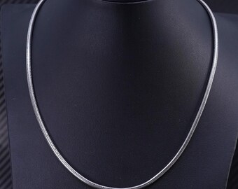 May include: A silver-colored necklace with a snake chain design. The necklace is displayed on a black mannequin, showcasing its sleek and simple style. The chain is thin and flexible, reflecting light.
