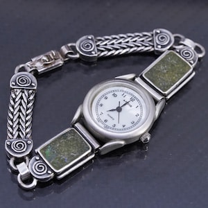 Puede incluir: Un reloj plateado con esfera blanca y números negros. La correa del reloj presenta un diseño tejido con detalles de piedra verde. La esfera del reloj dice "Annie Quartz". Un accesorio elegante.