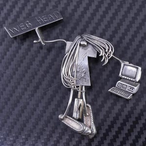 Puede incluir: Broche plateado con forma de persona sosteniendo un letrero que dice "WEB HEAD". La persona tiene pelo largo y sostiene una computadora. El broche tiene un diseño artístico único.