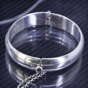 May include: A silver hinged bangle bracelet with a chain attached. The bracelet is smooth and shiny.