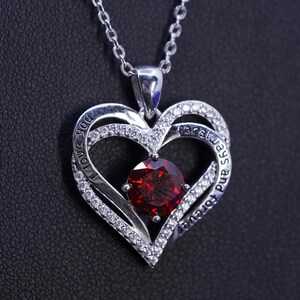 May include: A silver-toned heart pendant necklace with a large, round, red gemstone at the center. The pendant has a double-heart design, with the words "I love you" and "for always and forever" inscribed. The necklace chain is also silver.