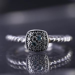 May include: A silver ring with a square, textured centerpiece. The centerpiece is encrusted with small, dark stones and a few blue stones. The band has a twisted rope design.