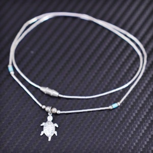 May include: A silver necklace with a small silver turtle pendant. The necklace is made of small silver beads and has a turquoise accent bead.